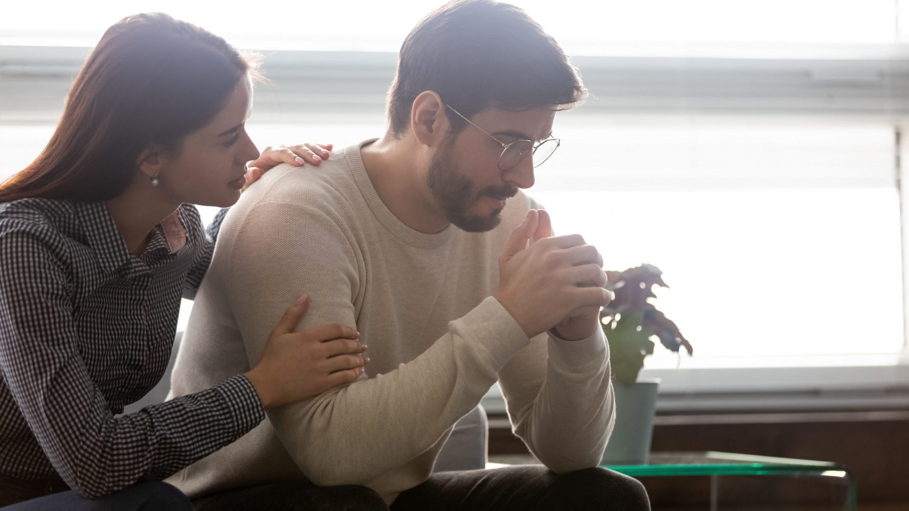 woman comforting man on couch in front of window.