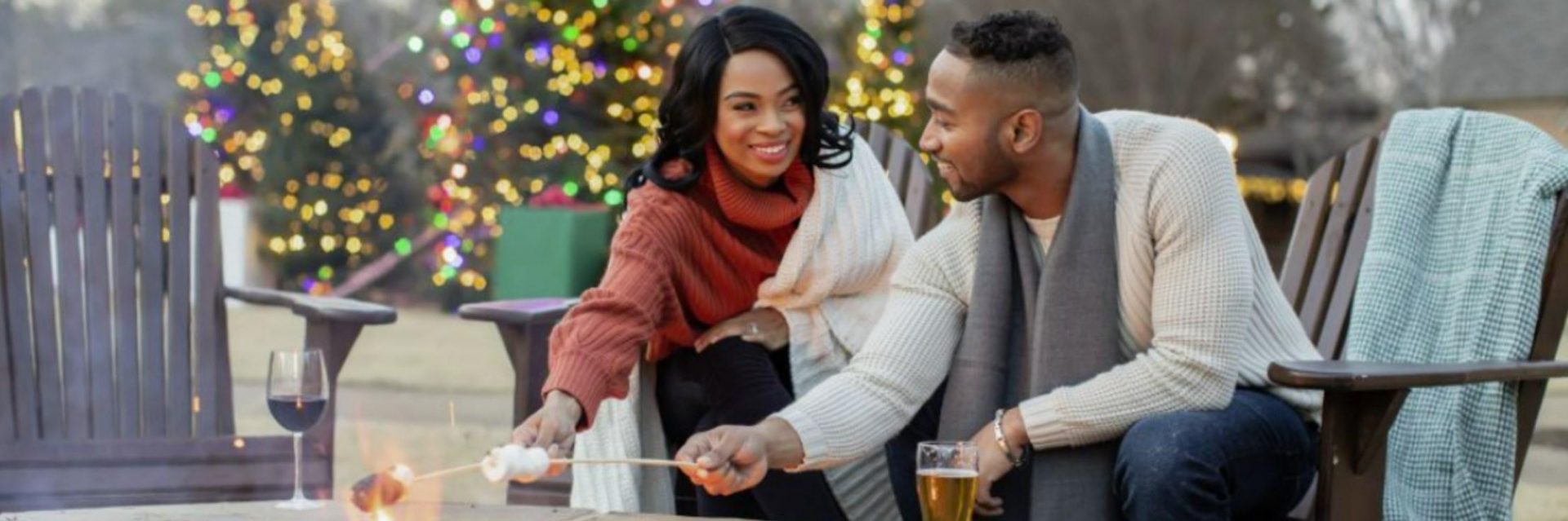 lovely couple sitting by the bonfire roasting marshmallows with christmas trees in the background.