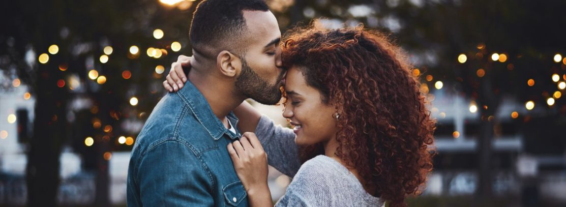 Boyfriend lovingly kissing girlfriend on forehead with trees in the background.