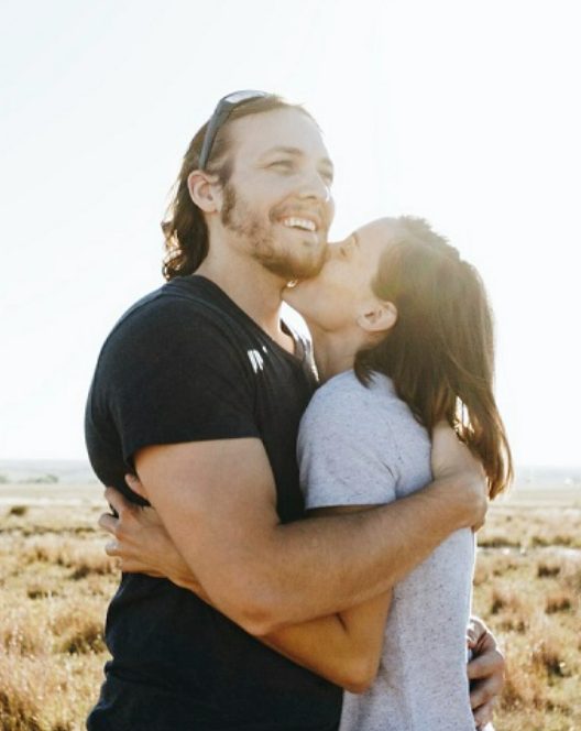 Girlfriend lovingly kissing her boyfriend's cheek in a field.