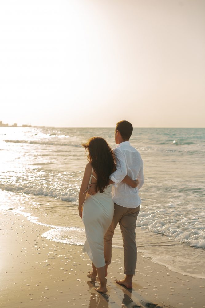 Boyfriend holding his girlfriend while walking down the beach with a beautiful sunset in the background. 