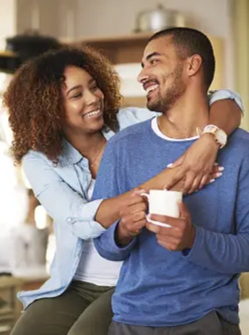 Girlfriend Hugging Boyfriend while they smile at each other lovingly. He is holding a coffee cup.