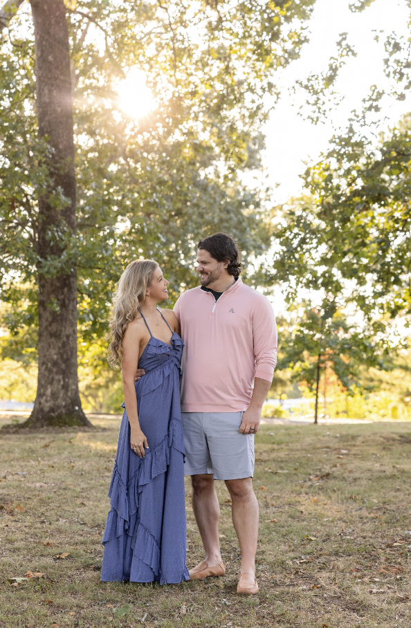boyfriend and girlfriend holding each other at the waist gazing at each other lovingly with the sun peeking in the background through the trees.