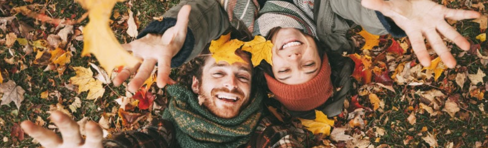 man and woman laying on the grass surrounded by fall leaves, throwing some up in the air.