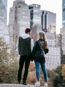 Male and Woman in their 30s looking at each other lovingly with the city skyline in the background. They are standing in a park.