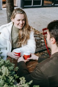 Couple on a coffee date. Woman is smiling warmly at the man sitting across from her. Both are holding red to-go coffee cups.