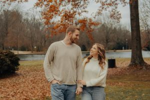 Couple looking lovingly each other under an autumn tree with orange leaves. They are both wearing cream colored sweaters.
