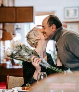 Elderly woman kissing her husband on the cheek as he hands her a bouquet of white roses.