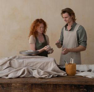 Red haired woman and male with mid length brown hair doing a pottery class together. They are both standing behind a table.